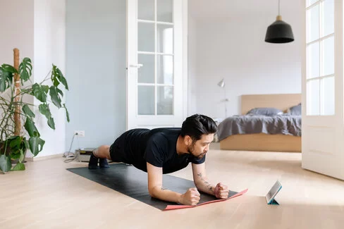 Ein Mann macht zu Hause Sport und trainiert im Unterarmstütz. Dabei schaut er auf ein Tablet.