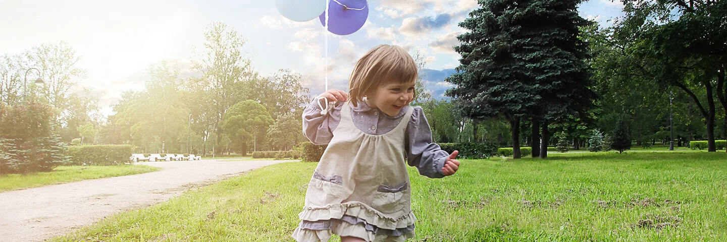 Ein kleines Mädchen läuft lachend über eine Wiese. In ihrer linken Hand hält sie einen Bündel Luftballons an einer Schnur.