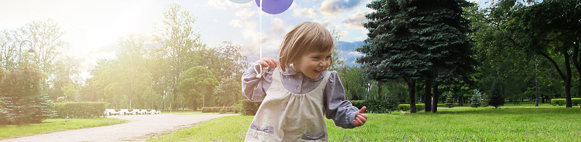 Ein kleines Mädchen läuft lachend über eine Wiese. In ihrer linken Hand hält sie einen Bündel Luftballons an einer Schnur.