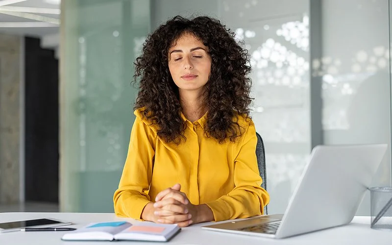 Eine Frau sitzt mit gefalteten Händen und geschlossenen Augen vor ihrem Laptop an ihrem Bürotisch.