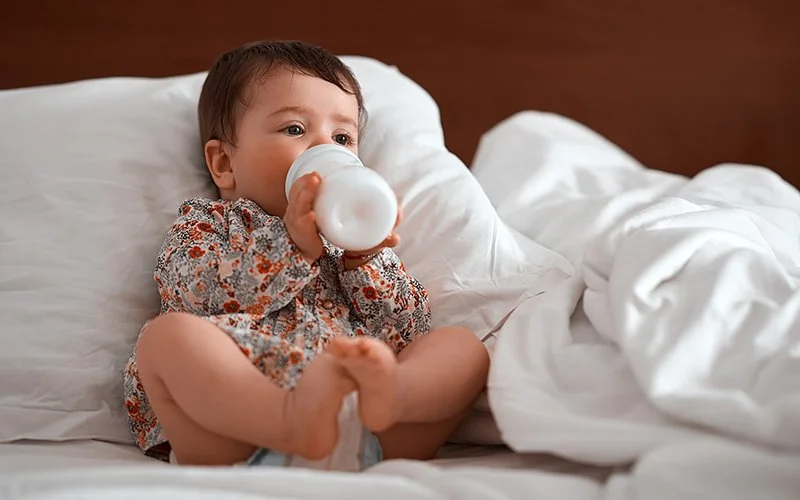 Ein Kleinkind sitzt auf einem Bett und trinkt aus einer Plastikflasche.