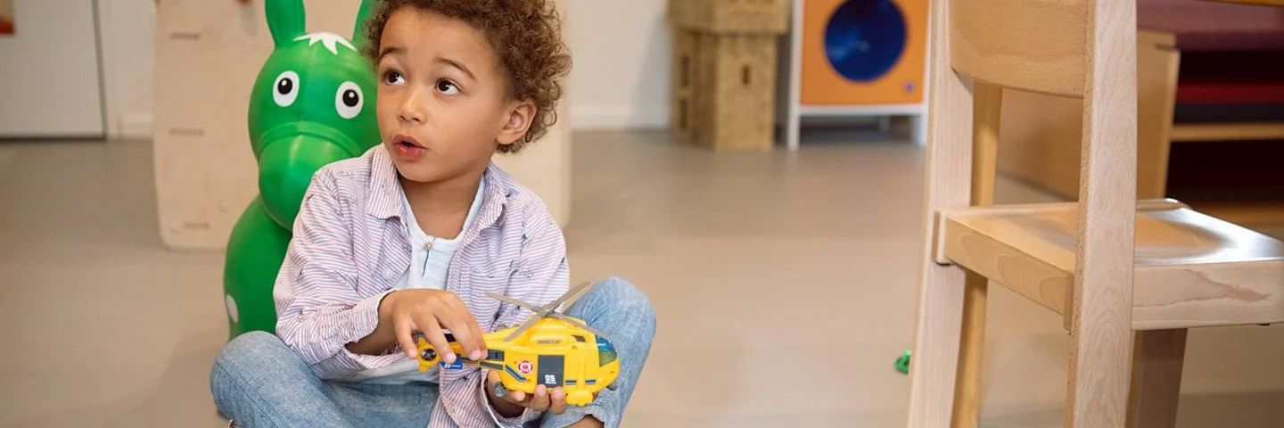 Ein Junge sitzt auf dem Boden eines Kindergartenzimmers und hält einen Spielzeughubschrauber in der Hand.