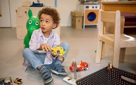 Ein Junge sitzt auf dem Boden eines Kindergartenzimmers und hält einen Spielzeughubschrauber in der Hand.