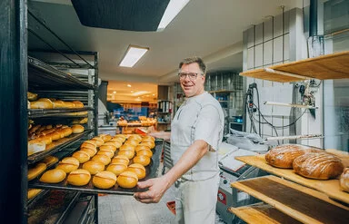 Der Bäckermeister Stefan Steeg schiebt in seiner Backstube ein großes Blech mit frisch gebackenen Brötchen in ein Backwarenregal.