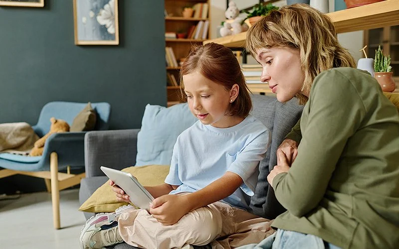 Eine Mutter sitzt mit ihrer Tochter im Grundschulalter auf einem Sofa und schaut, was ihre Tochter auf dem Tablet macht.