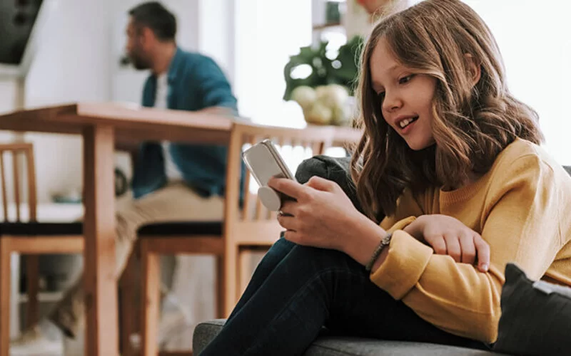 Ein junges Mädchen sitzt am Handy, der Vater im Hintergrund.