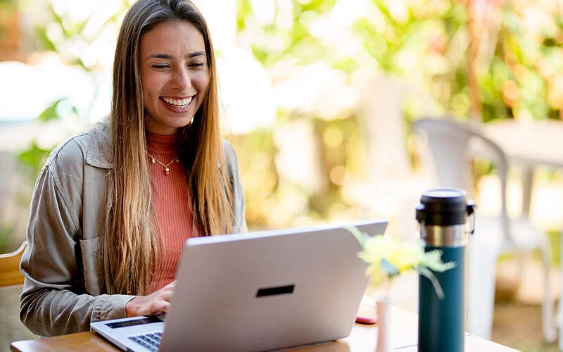 Eine junge Frau sitzt mit ihrem Laptop auf einer Terrasse und schaut lächelnd auf das Display.