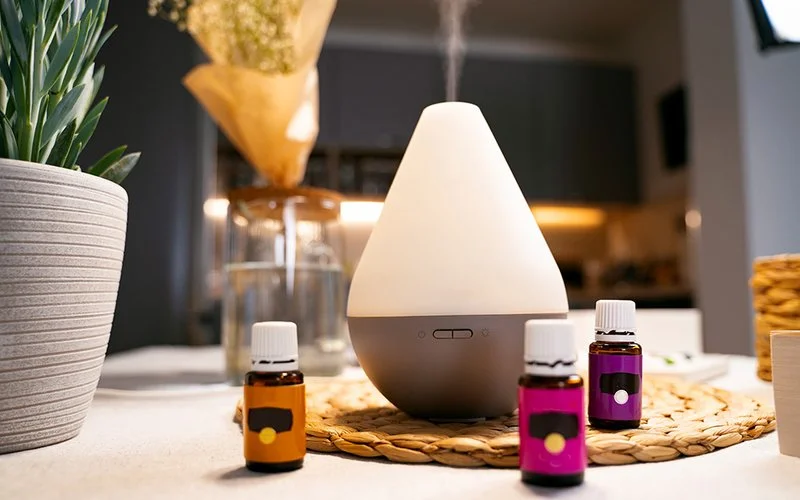 Auf einem Tisch stehen drei kleine braune Glasfläschchen mit ätherischen Ölen. Aus einem Diffusor strömt Dampf nach oben.