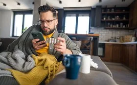 Ein Mann sitzt in Decken gewickelt auf einem Sofa. Er hält ein Smartphone in der einen und eine Tablettenpackung in der anderen Hand. Im Vordergrund steht eine blaue Tasse auf einem Tisch. Im Hintergrund sind eine offene Küche mit dunklen Holzschränken, weiße Fliesen und große Fenster zu sehen.