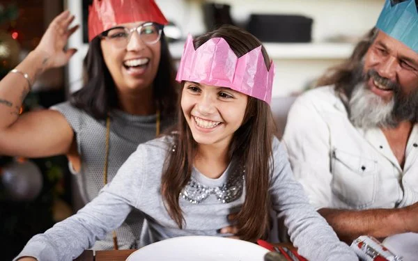 Kind mit pinker Papierkrone sitzt lachend am gedeckten Tisch; neben ihm Erwachsene mit Kronen, fröhliche Stimmung am Silvesterabend.