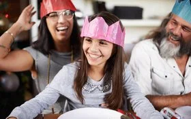 Kind mit pinker Papierkrone sitzt lachend am gedeckten Tisch; neben ihm Erwachsene mit Kronen, fröhliche Stimmung am Silvesterabend.