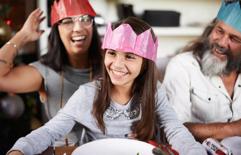 Kind mit pinker Papierkrone sitzt lachend am gedeckten Tisch; neben ihm Erwachsene mit Kronen, fröhliche Stimmung am Silvesterabend.