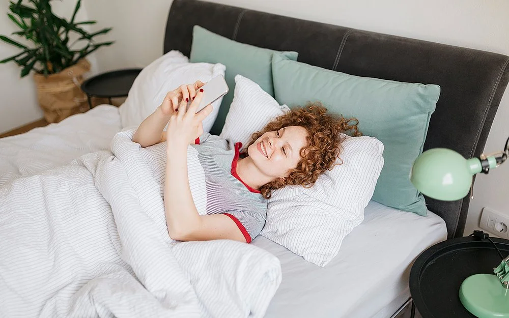 Eine junge Frau mit lockigen roten Haaren liegt im Bett und liest etwas auf ihrem Handy, dabei lächtelt sie.