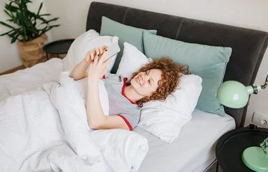 Eine junge Frau mit lockigen roten Haaren liegt im Bett und liest etwas auf ihrem Handy, dabei lächtelt sie.