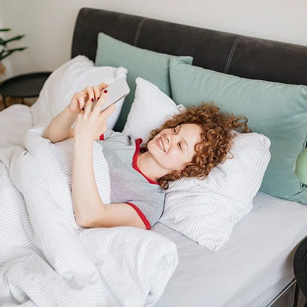 Eine junge Frau mit lockigen roten Haaren liegt im Bett und liest etwas auf ihrem Handy, dabei lächtelt sie.