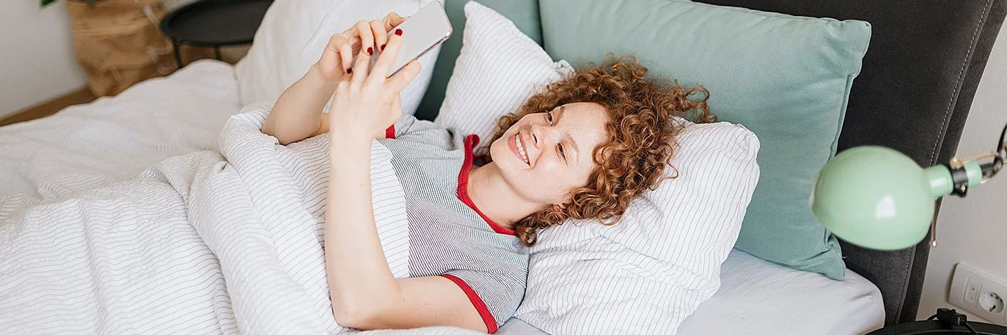 Eine junge Frau mit lockigen roten Haaren liegt im Bett und liest etwas auf ihrem Handy, dabei lächtelt sie.