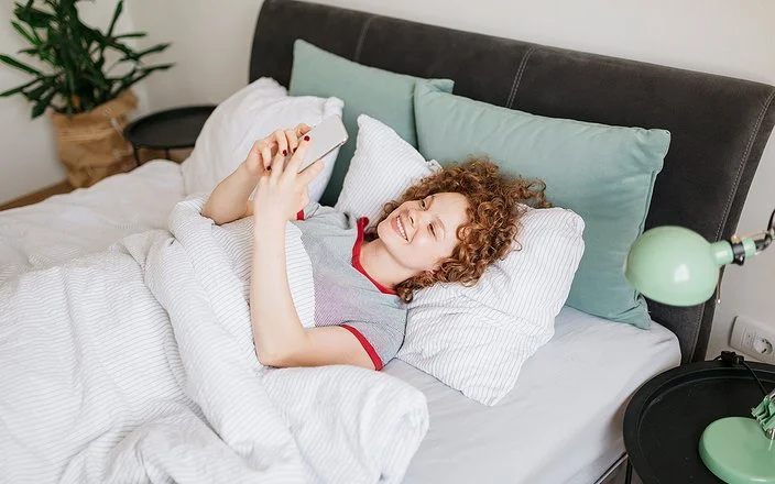 Eine junge Frau mit lockigen roten Haaren liegt im Bett und liest etwas auf ihrem Handy, dabei lächtelt sie.