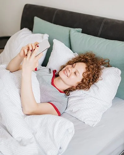 Eine junge Frau mit lockigen roten Haaren liegt im Bett und liest etwas auf ihrem Handy, dabei lächtelt sie.