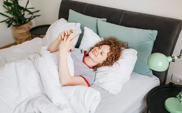 Eine junge Frau mit lockigen roten Haaren liegt im Bett und liest etwas auf ihrem Handy, dabei lächtelt sie.