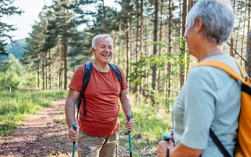 Ein etwa 60-jähriger Mann mit leichtem Bauchansatz auf einem Waldweg. Er trägt einen Rucksack und Walkingstöcke. Ihm gegenüber steht eine Frau, ebenfalls mit Walkingausrüstung. Beide scheinen in einem angeregten Gespräch. Der Mann lächelt die Frau an.