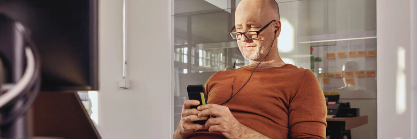 Ein Mann sitzt in einem Büro an einem Schreibtisch. Er schaut auf ein Smartphone, das er in den Händen hält.