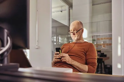 Ein Mann sitzt in einem Büro an einem Schreibtisch. Er schaut auf ein Smartphone, das er in den Händen hält.