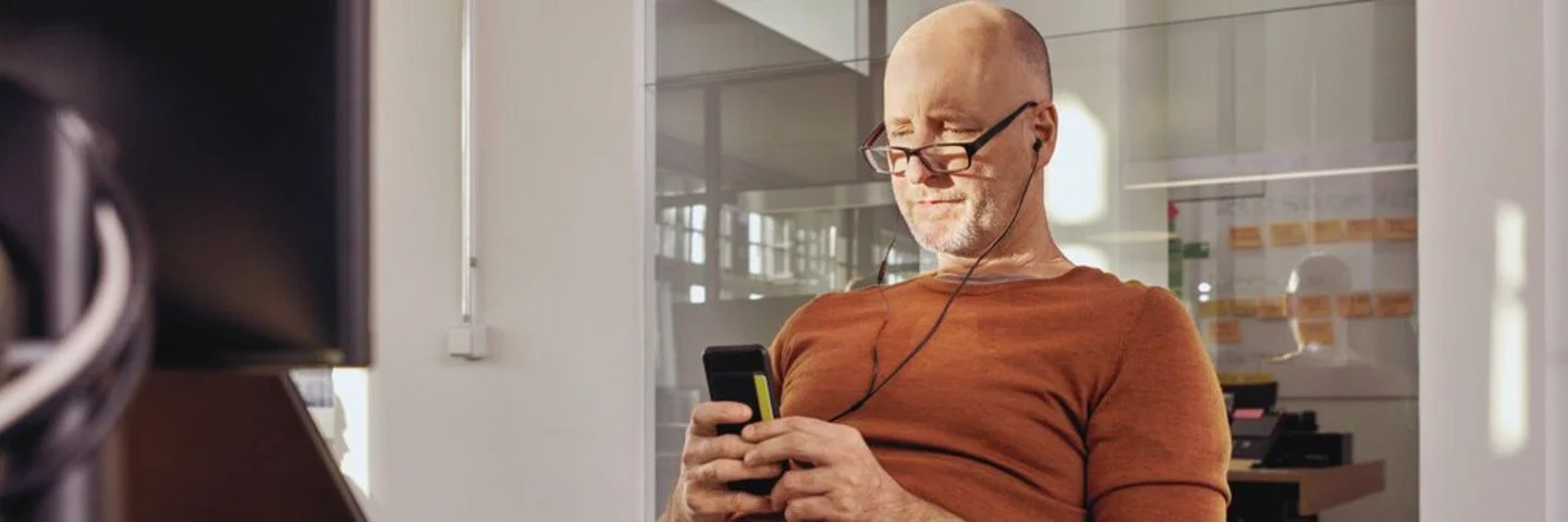 Ein Mann sitzt in einem Büro an einem Schreibtisch. Er schaut auf ein Smartphone, das er in den Händen hält.