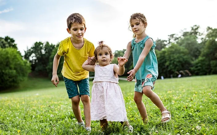 Zwei Kinder unterstützen ein kleines Mädchen bei den ersten Schritten auf einer grünen Wiese im Freien.