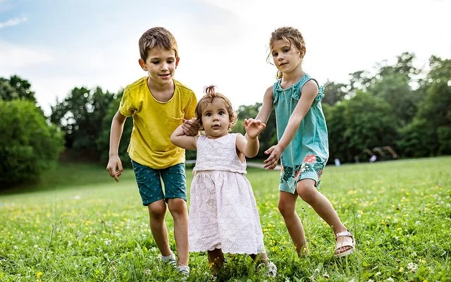 Zwei Kinder unterstützen ein kleines Mädchen bei den ersten Schritten auf einer grünen Wiese im Freien.