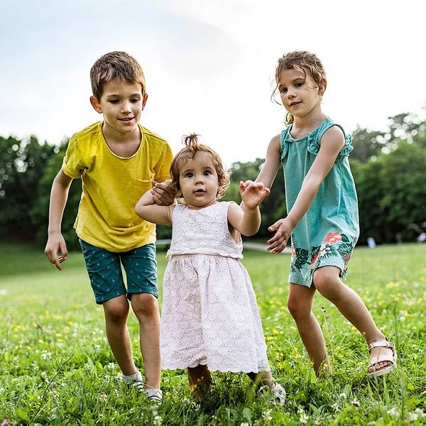 Zwei Kinder unterstützen ein kleines Mädchen bei den ersten Schritten auf einer grünen Wiese im Freien.