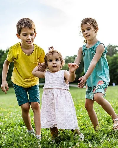 Zwei Kinder unterstützen ein kleines Mädchen bei den ersten Schritten auf einer grünen Wiese im Freien.