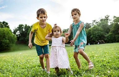 Zwei Kinder unterstützen ein kleines Mädchen bei den ersten Schritten auf einer grünen Wiese im Freien.