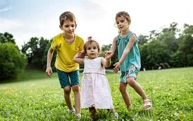 Zwei Kinder unterstützen ein kleines Mädchen bei den ersten Schritten auf einer grünen Wiese im Freien.