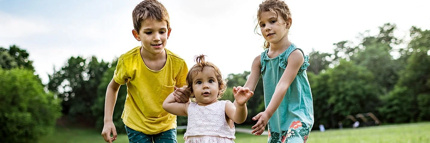 Zwei Kinder unterstützen ein kleines Mädchen bei den ersten Schritten auf einer grünen Wiese im Freien.
