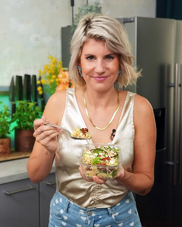 TV-Köchin Felicitas Then hält ein großes Glas mit Orzo-Salat in ihren Händen und präsentiert es freundlich lachend in die Kamera.