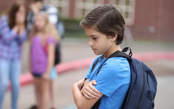 Ein Junge steht allein auf dem Schulhof und hält seine Arme vor die Brust. Im Hintergrund spielen Kinder.