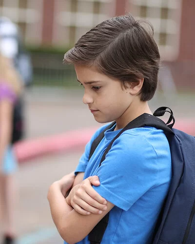 Ein Junge steht allein auf dem Schulhof und hält seine Arme vor die Brust. Im Hintergrund spielen Kinder.