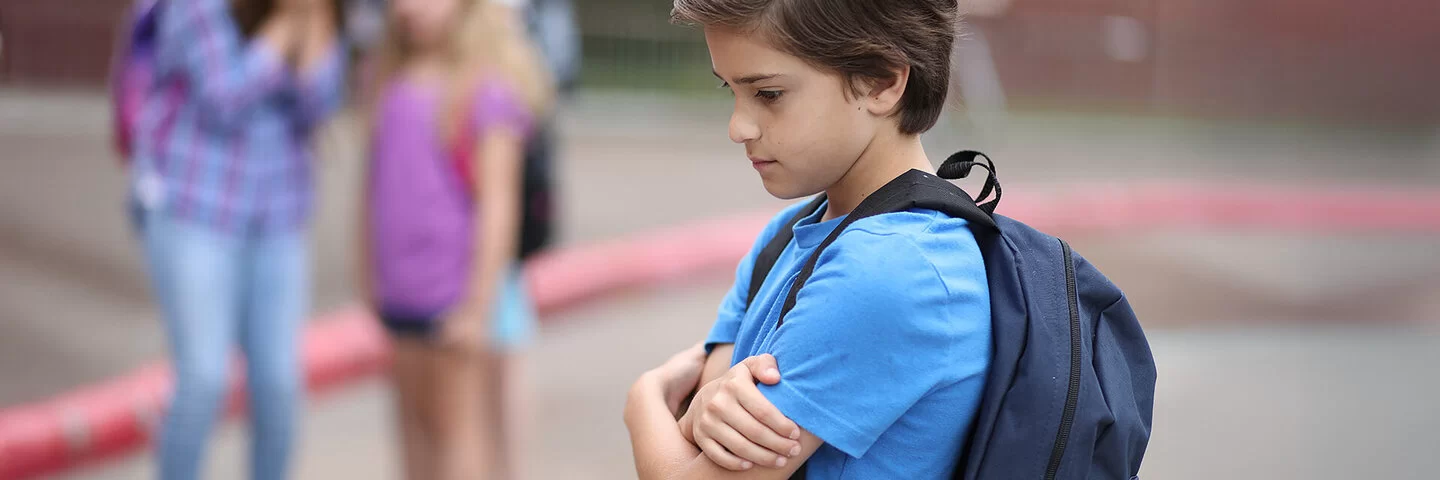 Ein Junge steht allein auf dem Schulhof und hält seine Arme vor die Brust. Im Hintergrund spielen Kinder.