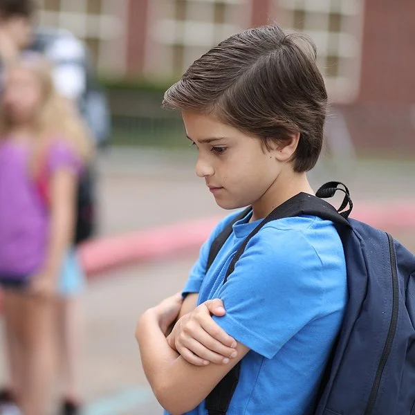 Ein Junge steht allein auf dem Schulhof und hält seine Arme vor die Brust. Im Hintergrund spielen Kinder.