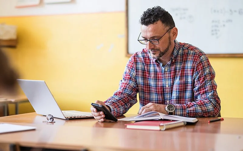 Ein Lehrer sitzt an seinem Pult und schaut auf sein Smartphone.
