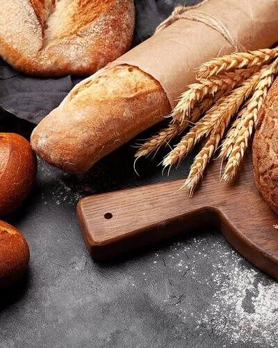 Verschiedene Weizenbackerzeugnisse auf einer Schieferplatte, darunter Brötchen, ein Baguette und ein angeschnittener Brotlaib auf einem Holzbrettchen. Zwischen Laib und Baguette liegt ein Bund Weizenähren.