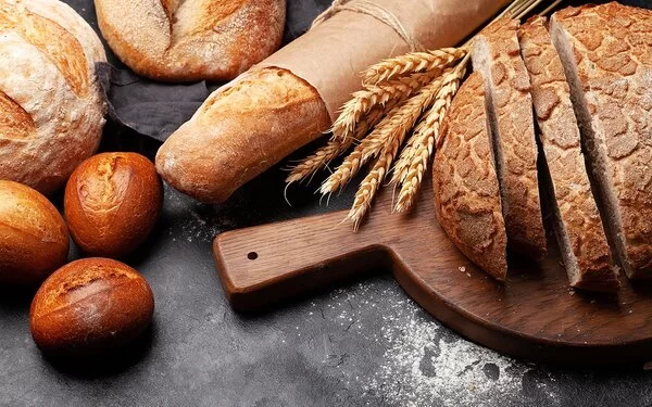 Verschiedene Weizenbackerzeugnisse auf einer Schieferplatte, darunter Brötchen, ein Baguette und ein angeschnittener Brotlaib auf einem Holzbrettchen. Zwischen Laib und Baguette liegt ein Bund Weizenähren.
