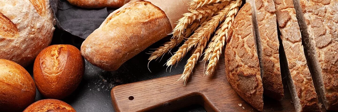 Verschiedene Weizenbackerzeugnisse auf einer Schieferplatte, darunter Brötchen, ein Baguette und ein angeschnittener Brotlaib auf einem Holzbrettchen. Zwischen Laib und Baguette liegt ein Bund Weizenähren.
