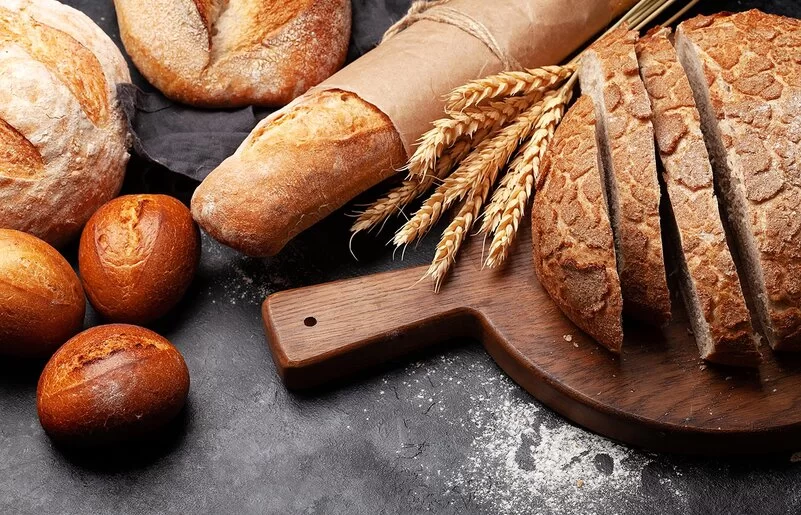 Verschiedene Weizenbackerzeugnisse auf einer Schieferplatte, darunter Brötchen, ein Baguette und ein angeschnittener Brotlaib auf einem Holzbrettchen. Zwischen Laib und Baguette liegt ein Bund Weizenähren.