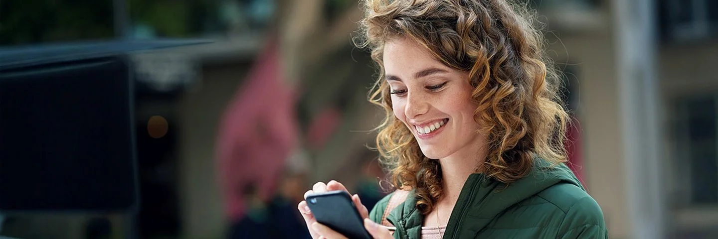 Eine junge lächelnde Frau in einer grünen Jacke schaut auf ihr Smartphone und tippt auf den Bildschirm.