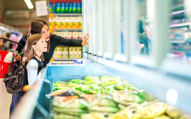 Mutter steht mit Tochter vorm Tiefkühlregal im Supermarkt.