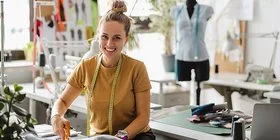 Junge Modedesignerin steht mit einer Schere bei der Arbeit an ihrem Arbeitsplatz und schneidet Stoff.