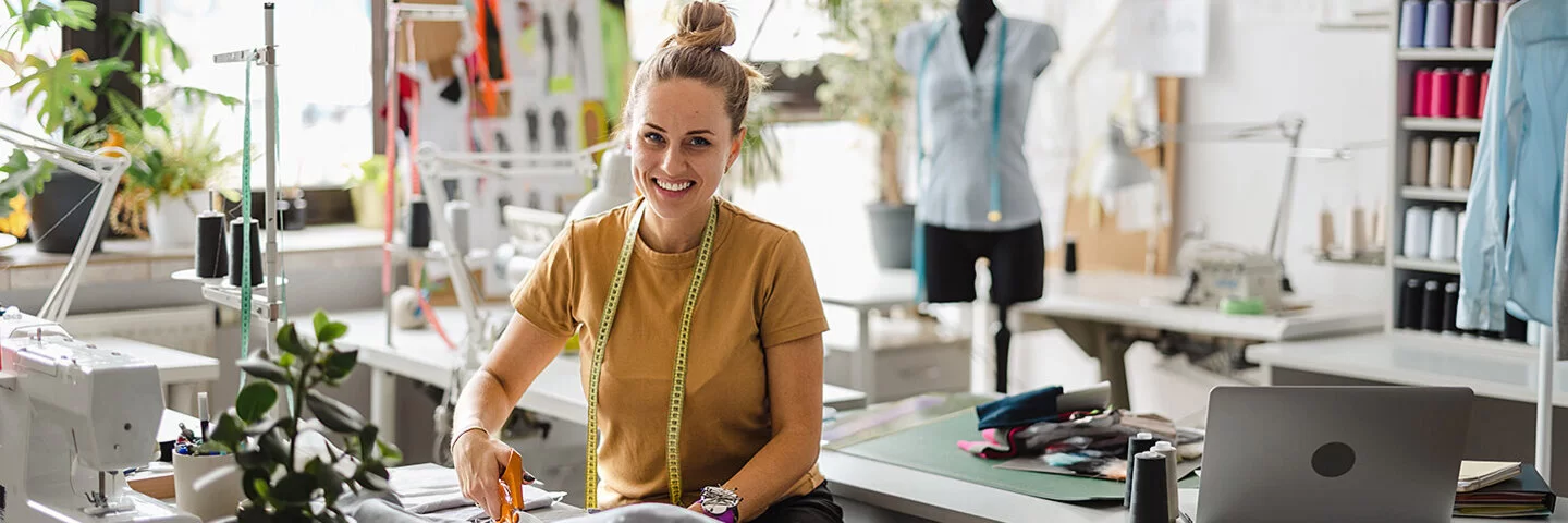 Junge Modedesignerin steht mit einer Schere bei der Arbeit an ihrem Arbeitsplatz und schneidet Stoff.