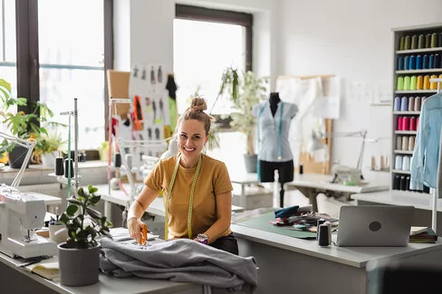 Junge Modedesignerin steht mit einer Schere bei der Arbeit an ihrem Arbeitsplatz und schneidet Stoff.