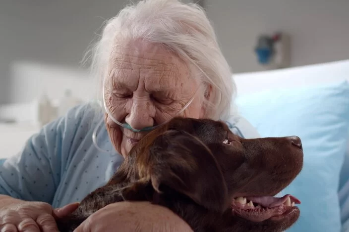 Eine ältere Frau wird über einen Schlauch in der Nase mit Sauerstoff beatmet. Sie umarmt einen Hund.
